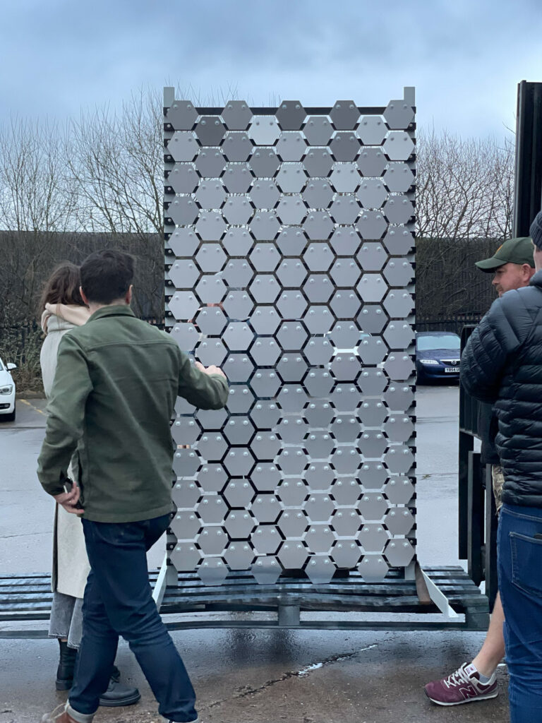 Members of the SEE MONSTER team survey a panel of Monster scales - a series of hexagonal aluminium tiles mounted side by side.