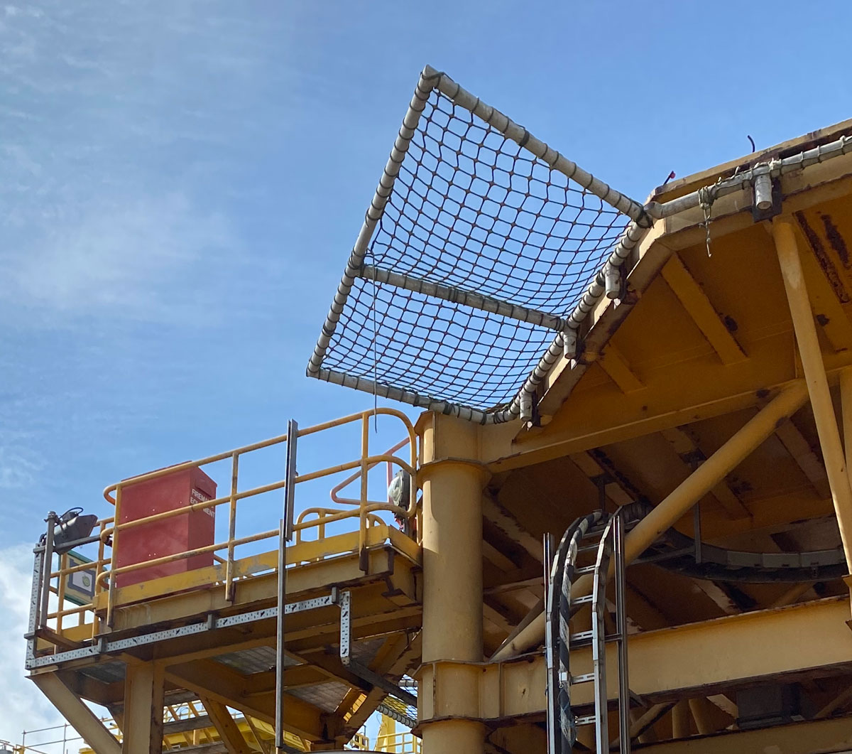 Looking up at the corner of an offshore platform, with yellow steel bars criss-crossing and a safety net stretching up and out from the structure. The sky is blue behind.