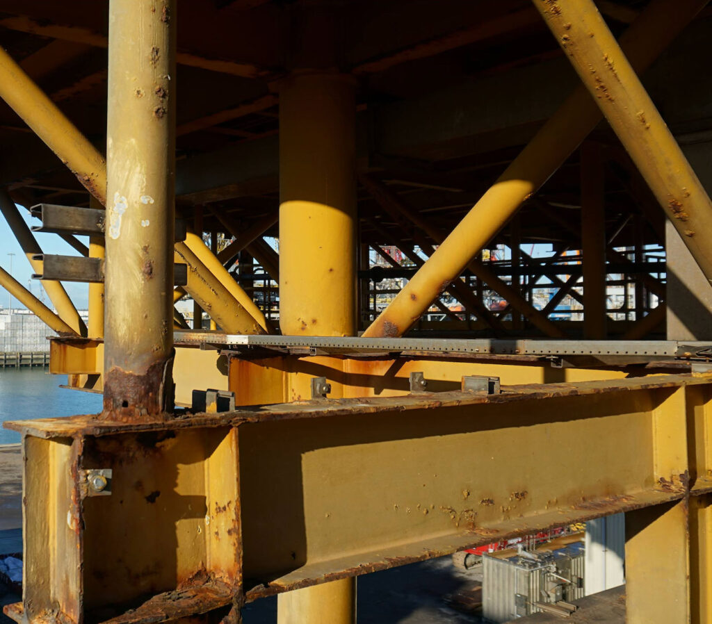 The rusted yellow steel beams of an offshore platform criss-cross in the foreground. In the background, shipping containers are partially visible in an industrial waterside setting.